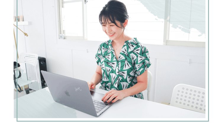 白い部屋でノートパソコンに向かい微笑む緑柄トップス姿の女性
