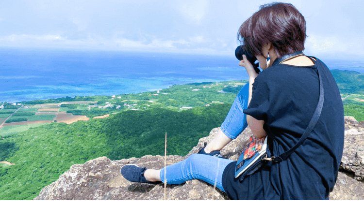岩場の崖に座り、カメラで海と緑の丘を撮影する後ろ姿の小山志織さん