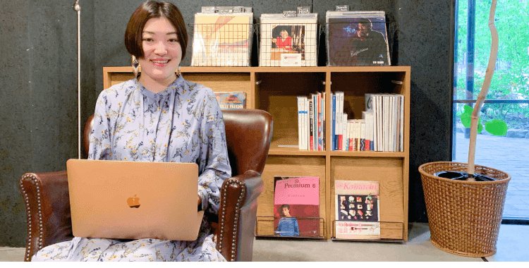 本棚のある部屋でひじ掛け椅子に座り、ノートPCに向かい微笑む小山志織さん