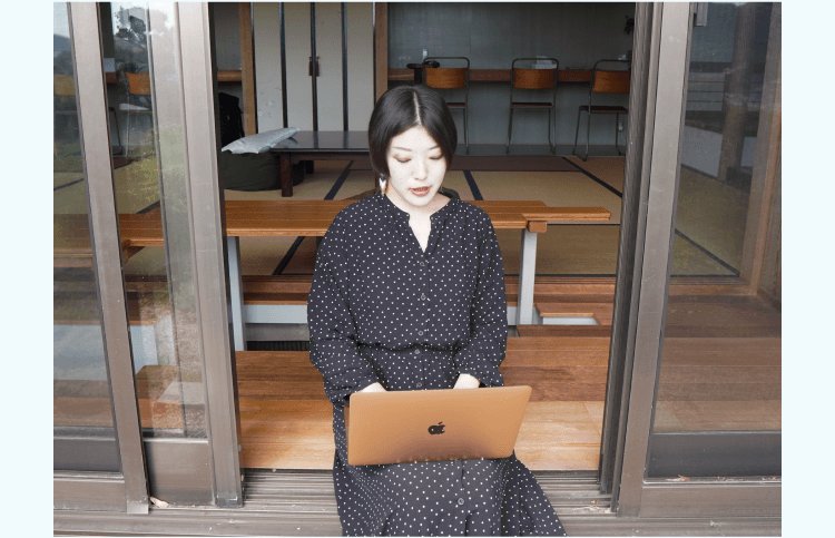 縁側でノートPCに向かい微笑む小山志織さん(ドット柄ワンピース)