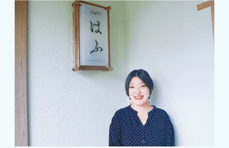 「HafHはふ」と書かれた木製の看板が壁に掛かった部屋の前で微笑む小山志織さん