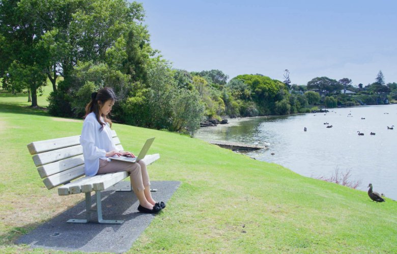 芝生の公園のベンチに座り、湖を眺める鴨を横目にノートパソコンを操作するちひろさんの写真