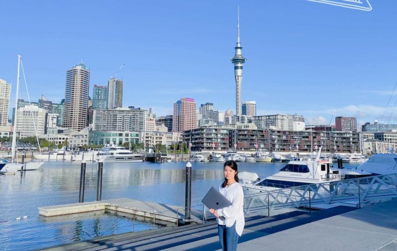 オークランドの港・高層ビル群・スカイタワーを背景に、ノートパソコンを抱えて桟橋に立つちひろさんの写真