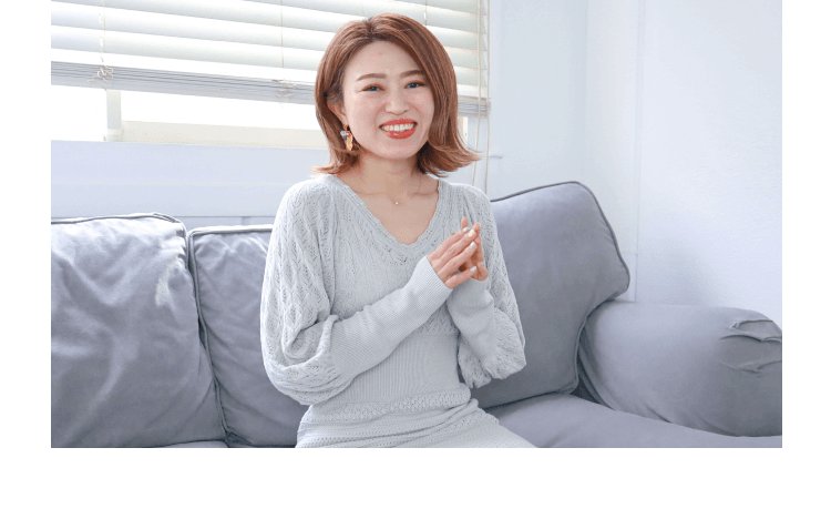 グレーのソファに座り、胸に手を当てて微笑む女性