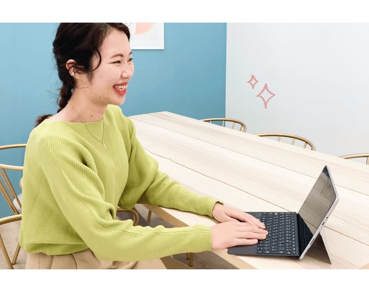 木製の長テーブルでノートパソコンを操作しながら微笑む黄緑色のトップスのシホさん