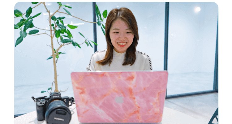 ピンクの大理石調ノートPCに向かうかんなさん（観葉植物、カメラが机に置かれている、窓際）