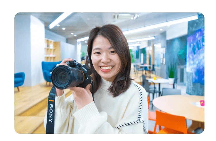 笑顔でカメラを構えるかんなさん（SONYストラップのカメラ、コワーキングスペース内）