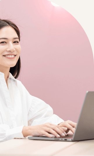白いブラウスの石井さんがピンクのアーチ装飾を背にノートPCに向かい微笑む写真