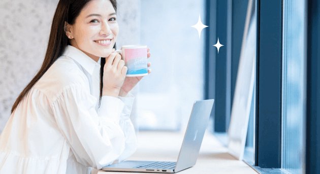 窓辺でマグカップを持ちノートPCの前で微笑む石井さんの写真