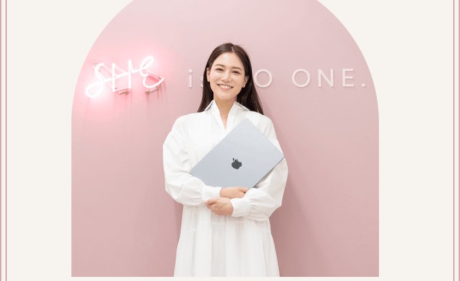 ピンクのアーチ壁と「SHE is NO ONE.」のネオンサインを背に、ノートPCを抱えて立つ石井さんの写真