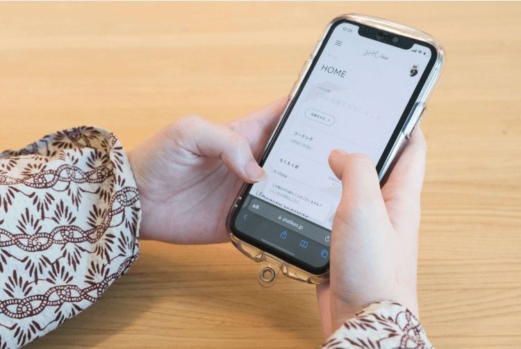 木製テーブルの上でスマートフォンのSHElikesアプリ画面を操作する手元
