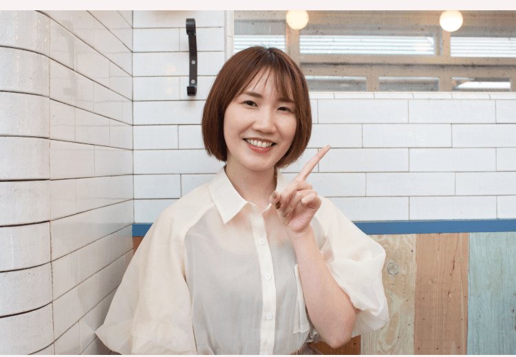 タイル壁を背に指を立てて話すかのりさん