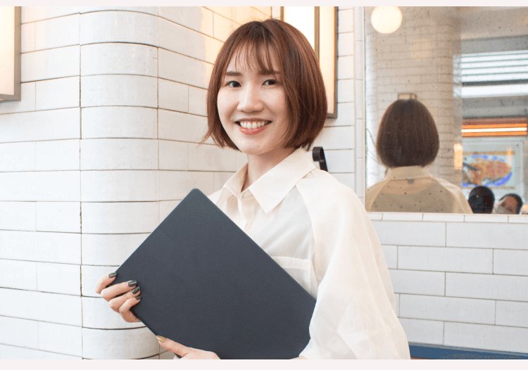 タイル壁を背にファイルを抱えて微笑むかのりさん