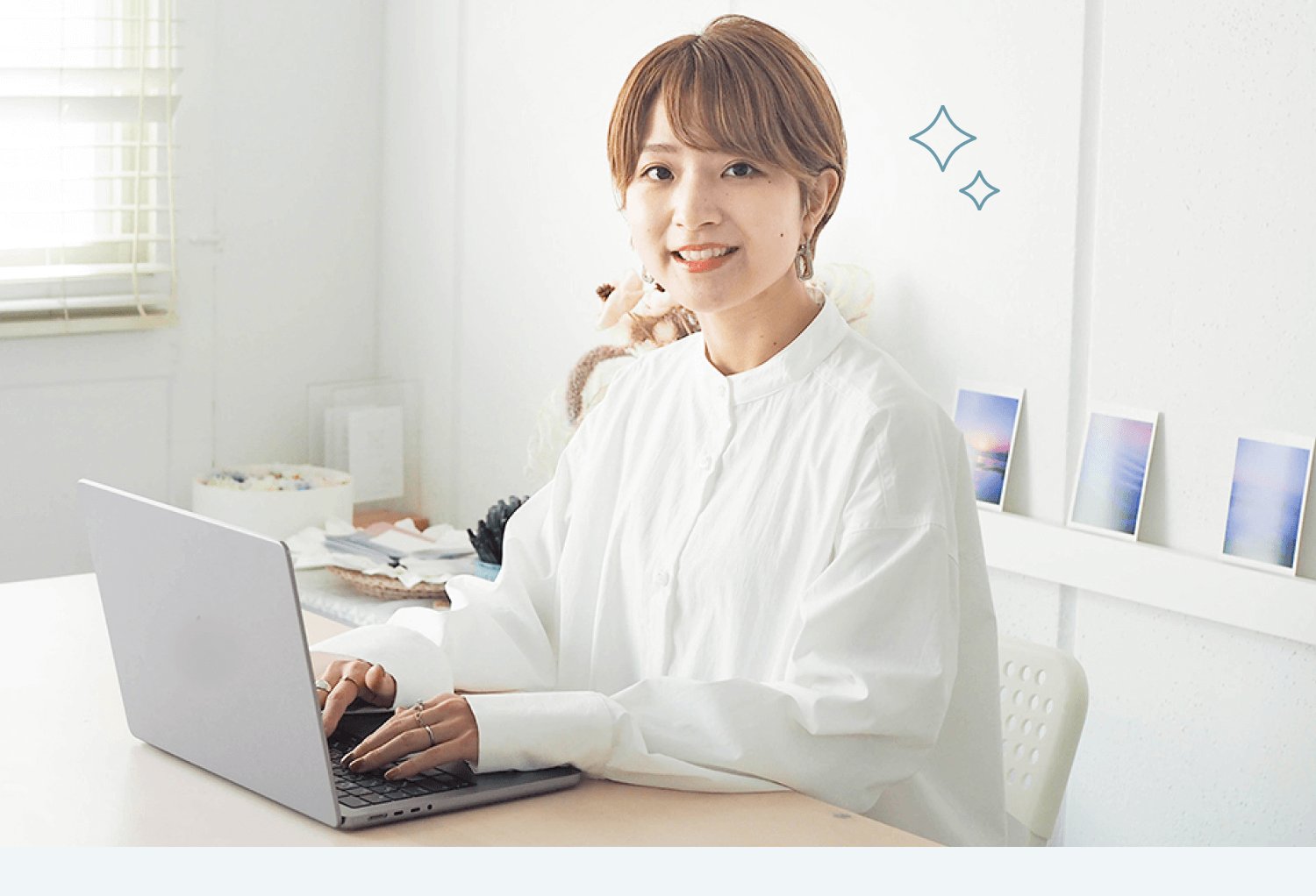 白いブラインドの部屋でノートPCに向かい微笑む白シャツの女性
