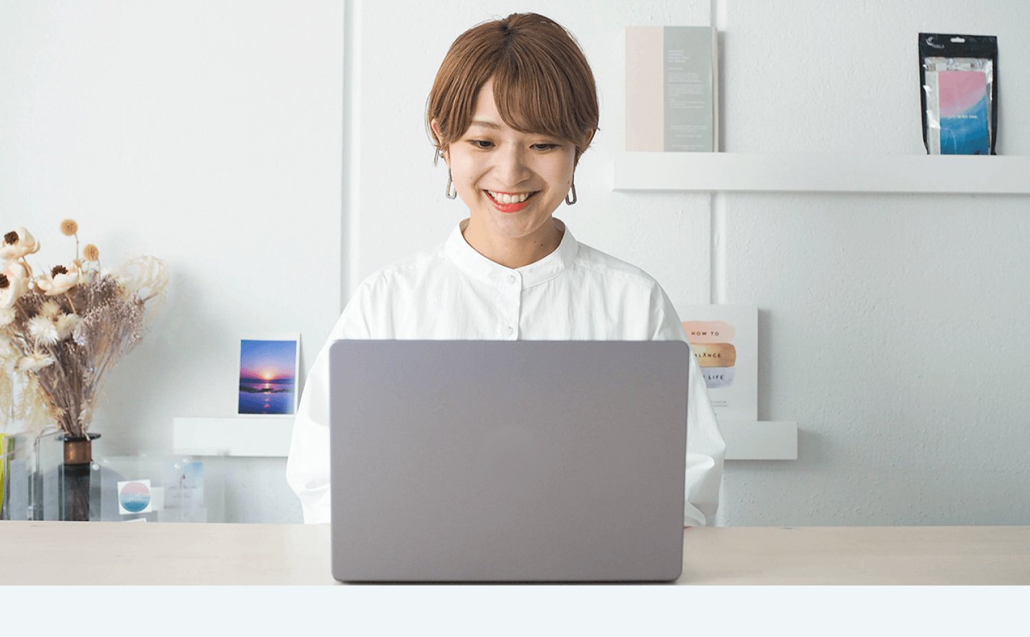 ドライフラワーのそばでノートPCに向かい微笑む白シャツの女性