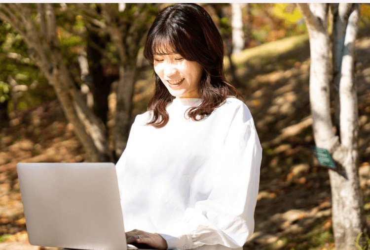 紅葉の林で微笑みながらノートPCを操作する女性(まみこさん)