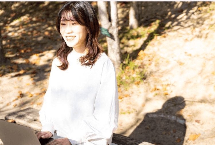 紅葉の中、木のベンチでノートPCを操作しながら微笑む女性(まみこさん)