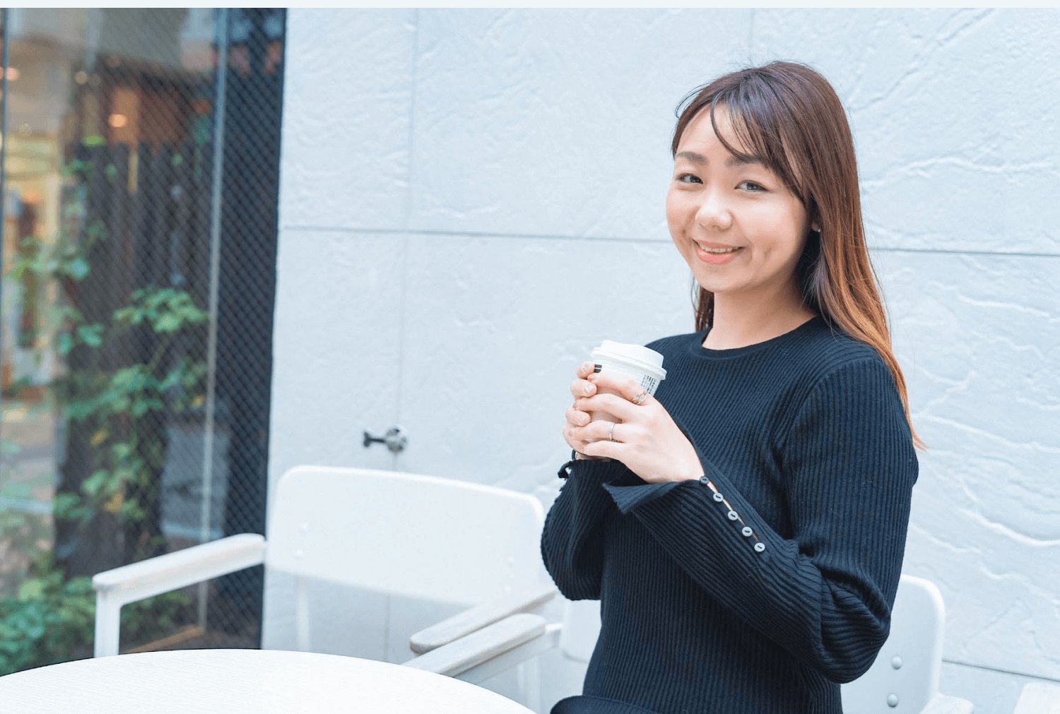 紺のニットでコーヒーカップを持って立つReinaさん（屋外・白タイル壁）