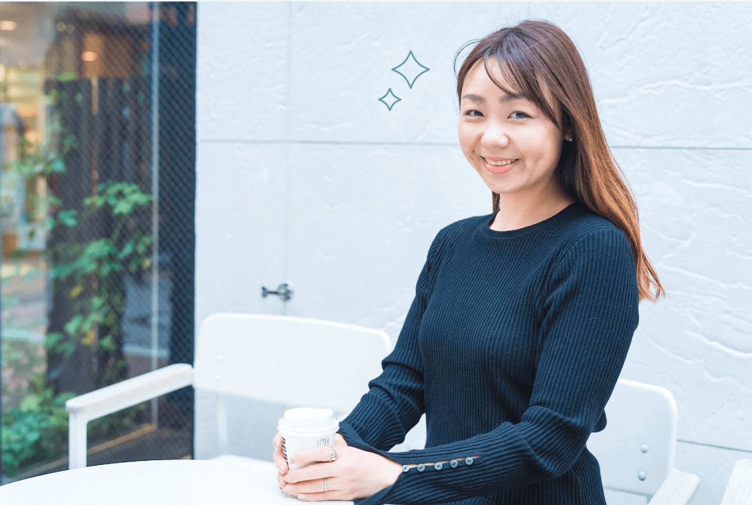 紺のニットでコーヒーカップを持って着席するReinaさん（屋外・白タイル壁）