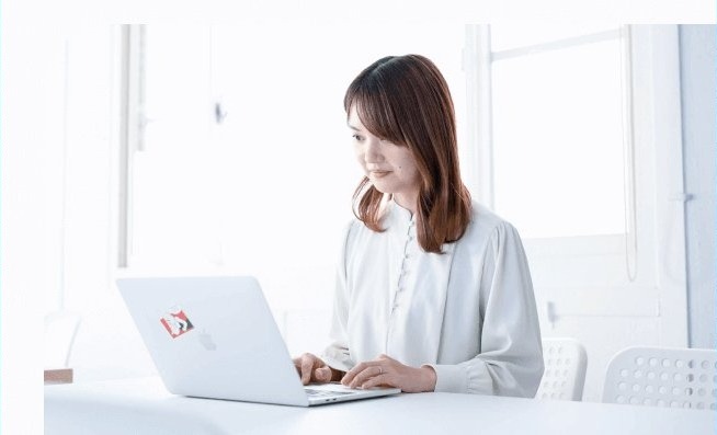 白いテーブルでノートPCに向かう女性、窓辺の明るい室内