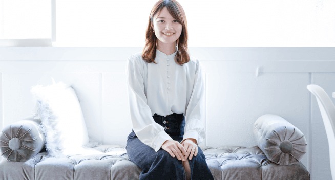 グレーのソファに腰掛け、クッションに囲まれて微笑む女性