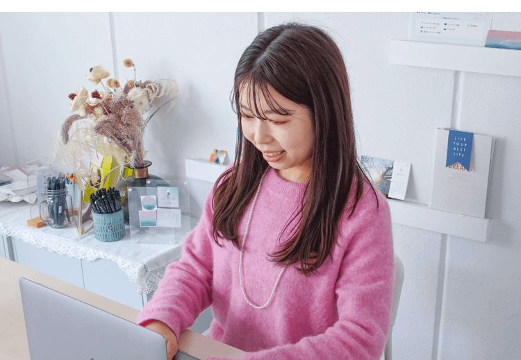 自宅デスクでノートパソコンに向かうyurieさん（花・雑貨の棚が背景）