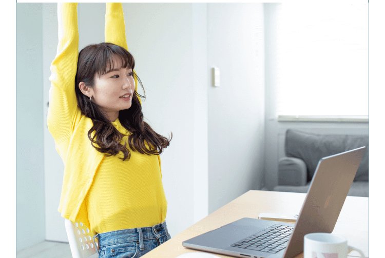 両手を上げてストレッチする黄色トップスの女性、奥の机にノートPC