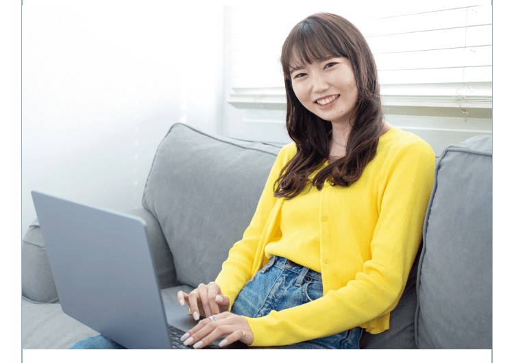 ソファでノートPCを操作しながら微笑む黄色カーディガンの女性