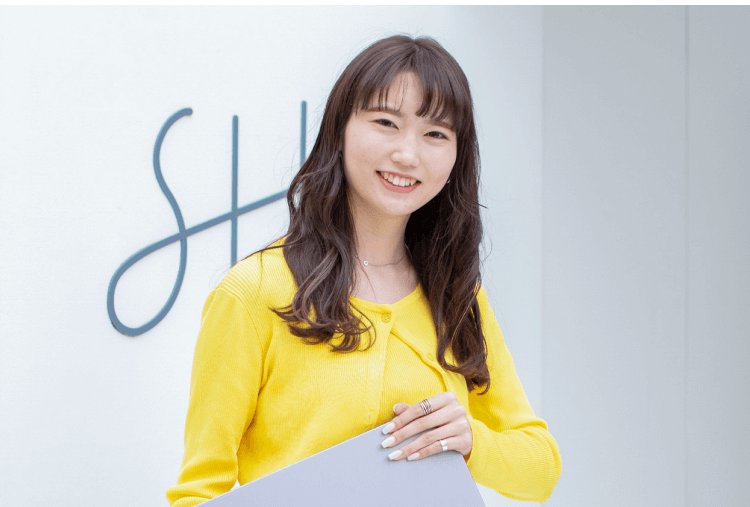 SHElikesのロゴサイン脇に立ち微笑む黄色カーディガンの女性