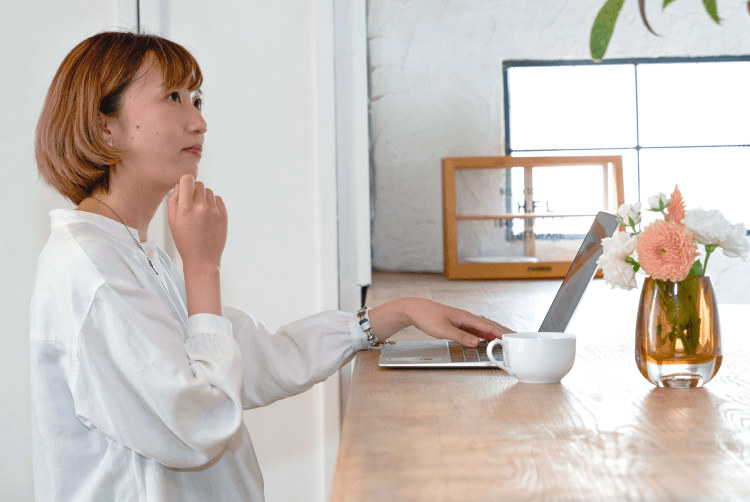 白いトップスでノートPCに手を伸ばしながら考え込む表情のろこさん。窓際のカウンターに花瓶とコーヒーカップ