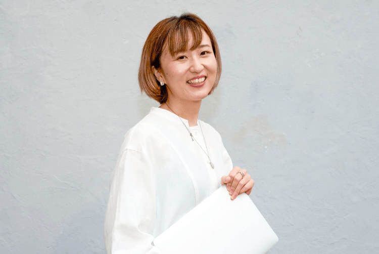 白いトップスで微笑むろこさんのポートレート。灰色の壁を背景に白いファイルを抱えている