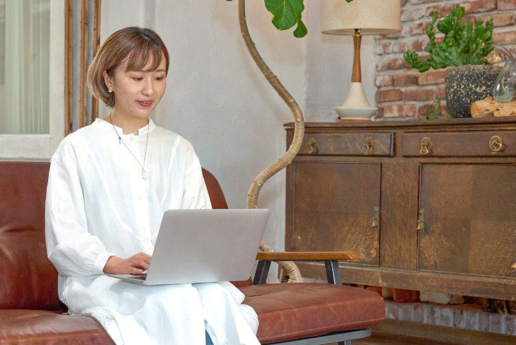 アンティークなソファに座り、ノートPCを操作しながら話すろこさん。背景にレンガ壁・観葉植物・木製キャビネット