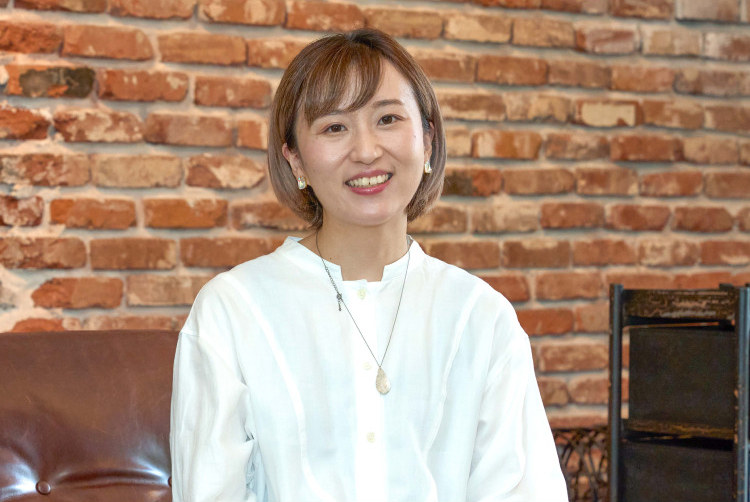 レンガ壁を背景に微笑むろこさんのポートレート。白いブラウス姿で椅子に座っている