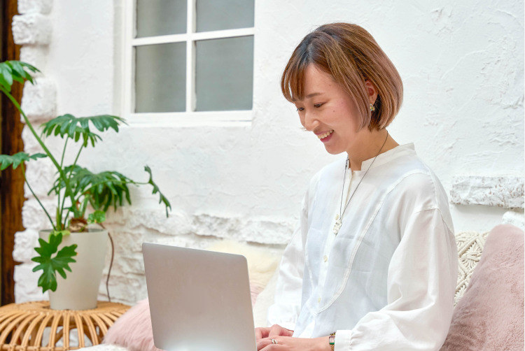 観葉植物と窓辺のあるスペースで、ノートPCを見ながら微笑むろこさんのポートレート