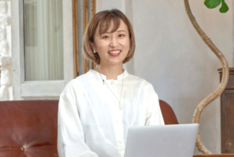 ソファに座り、ノートPCの前で微笑むろこさんのポートレート。背景にレンガ壁・観葉植物・木製キャビネット