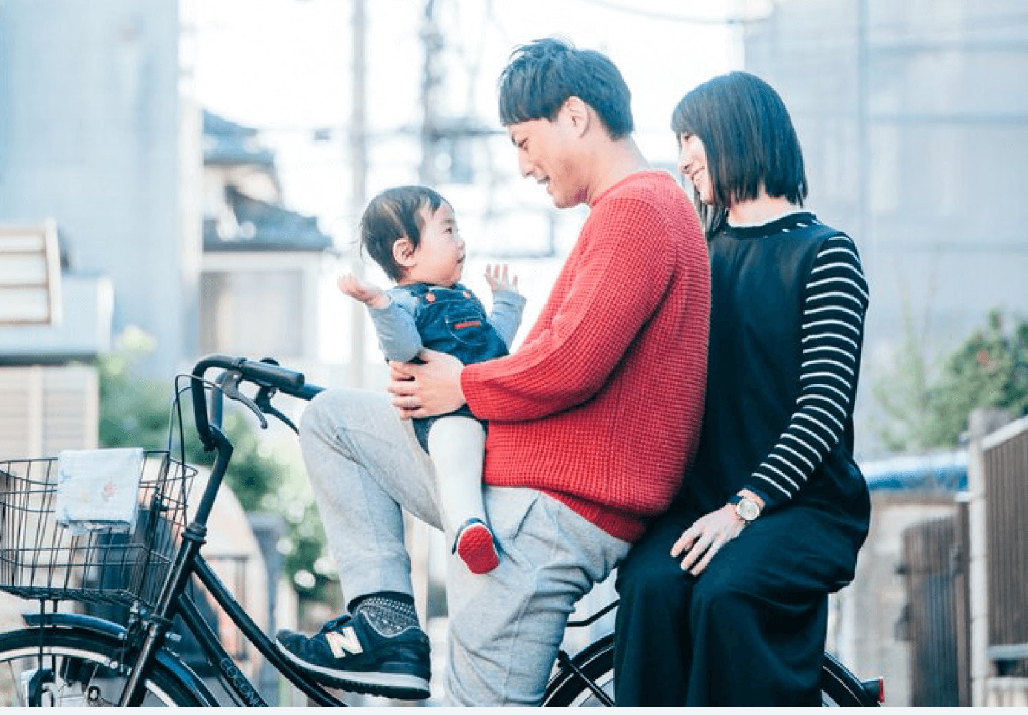 自転車にまたがり赤ちゃんを抱く夫の後ろに横座りする、きゃんまりさんの3人家族の屋外スナップ
