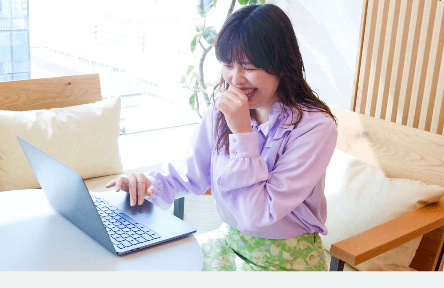 白いソファに座り、ノートPCを操作しながら笑う女性
