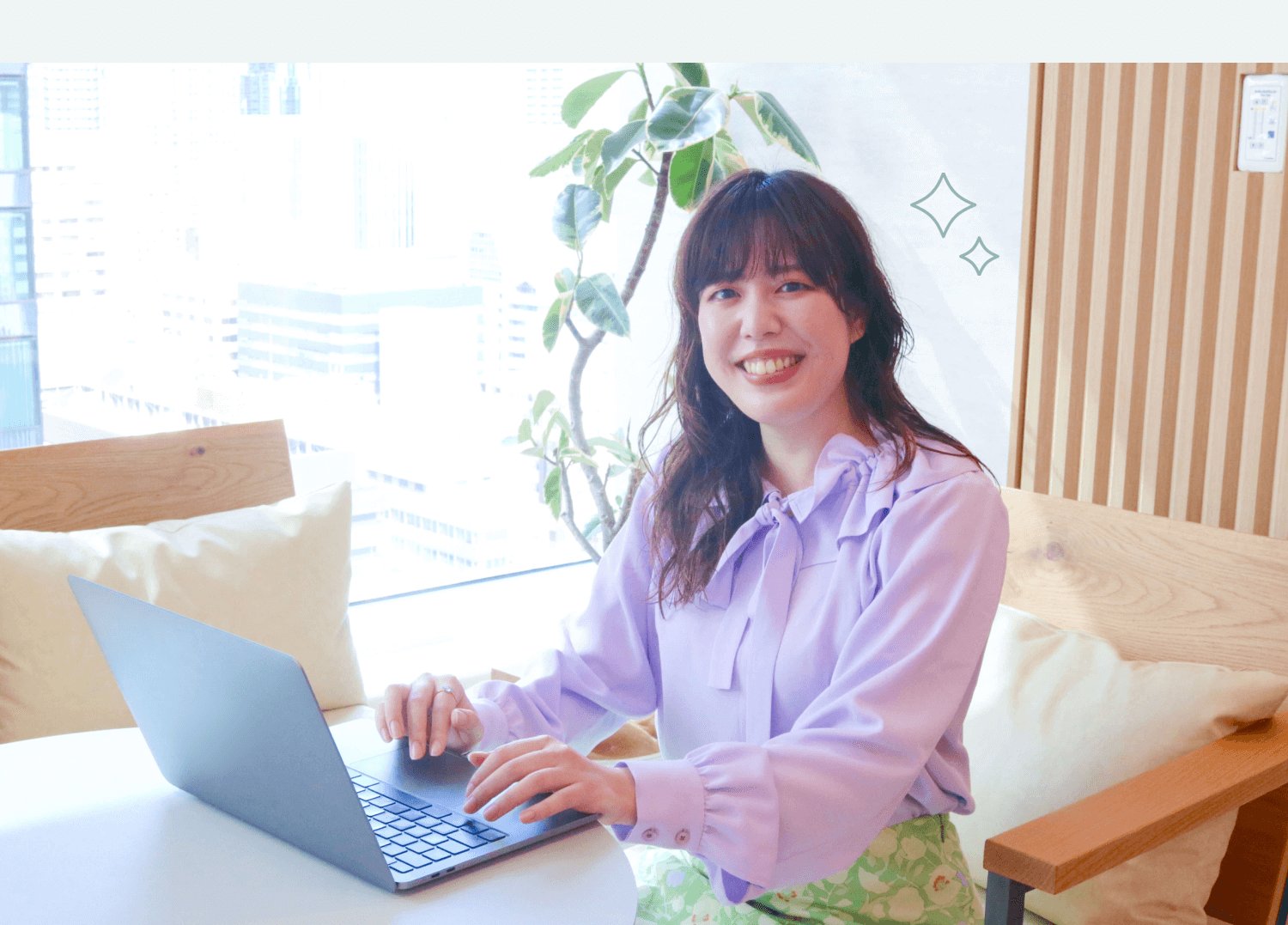 観葉植物と窓辺の明るい室内で、ノートPCを操作しながら微笑む女性