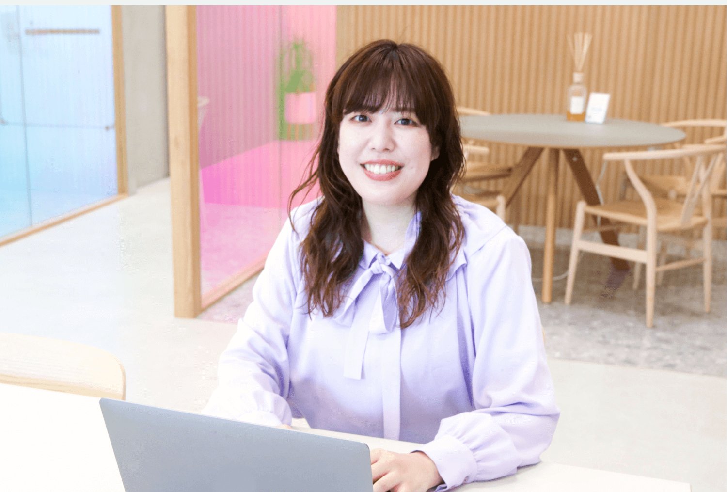 ピンク・ブルーのガラスパーティションが見えるオフィス空間で、ノートPCを抱えて微笑む女性