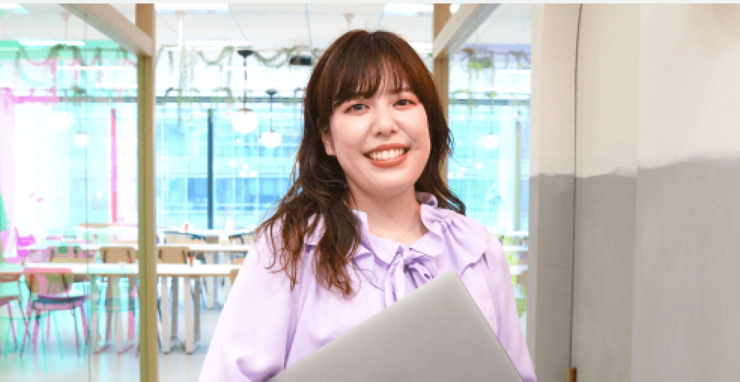 ガラス張りのオフィスの通路で、ノートPCを抱えて微笑む女性