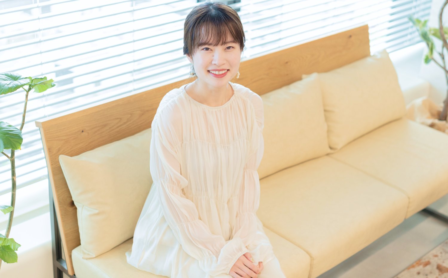 窓辺のソファに座り微笑む女性（観葉植物・ブラインド背景）