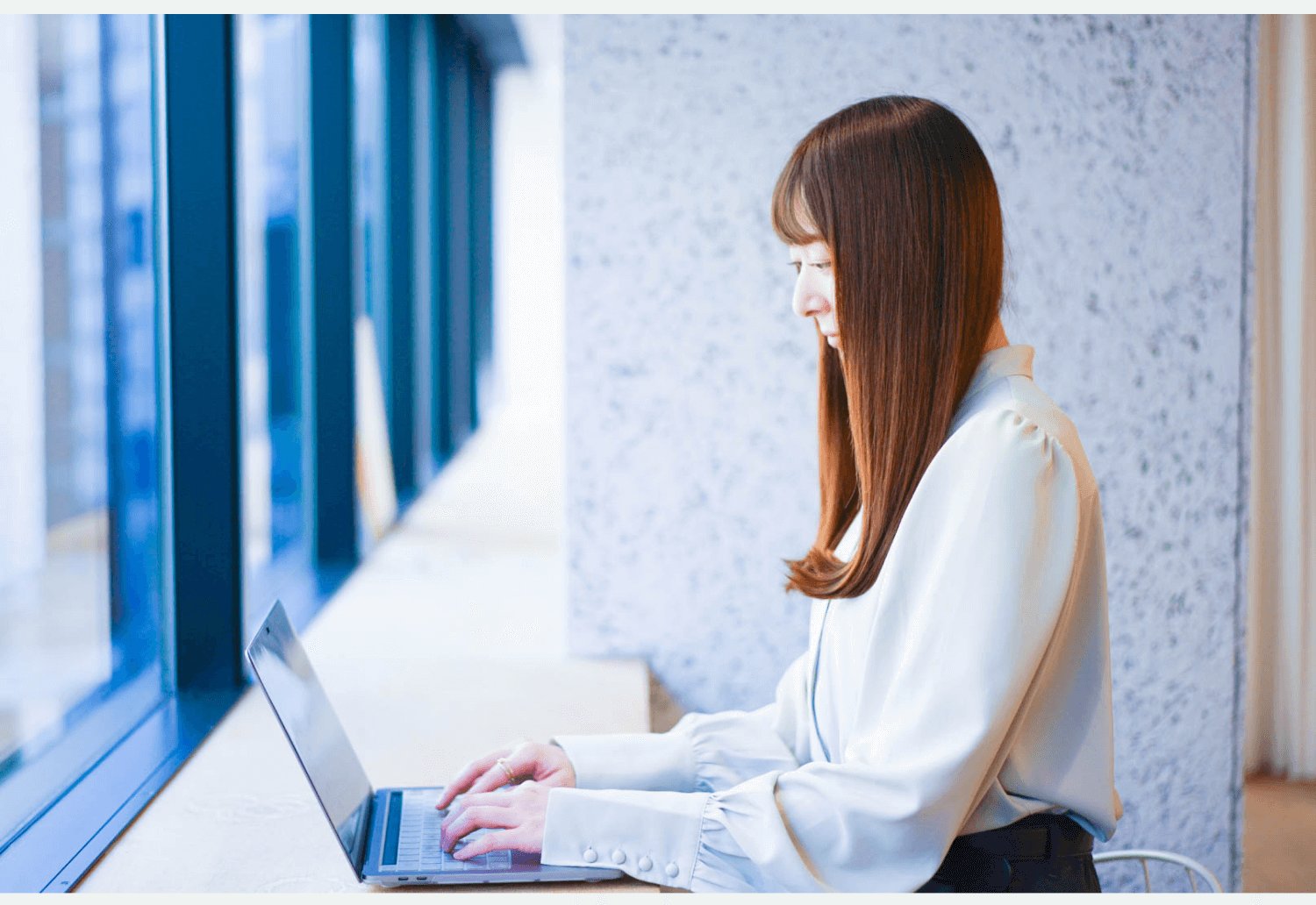 木目の壁とガラス窓を背に、テーブルでノートPCを操作する女性