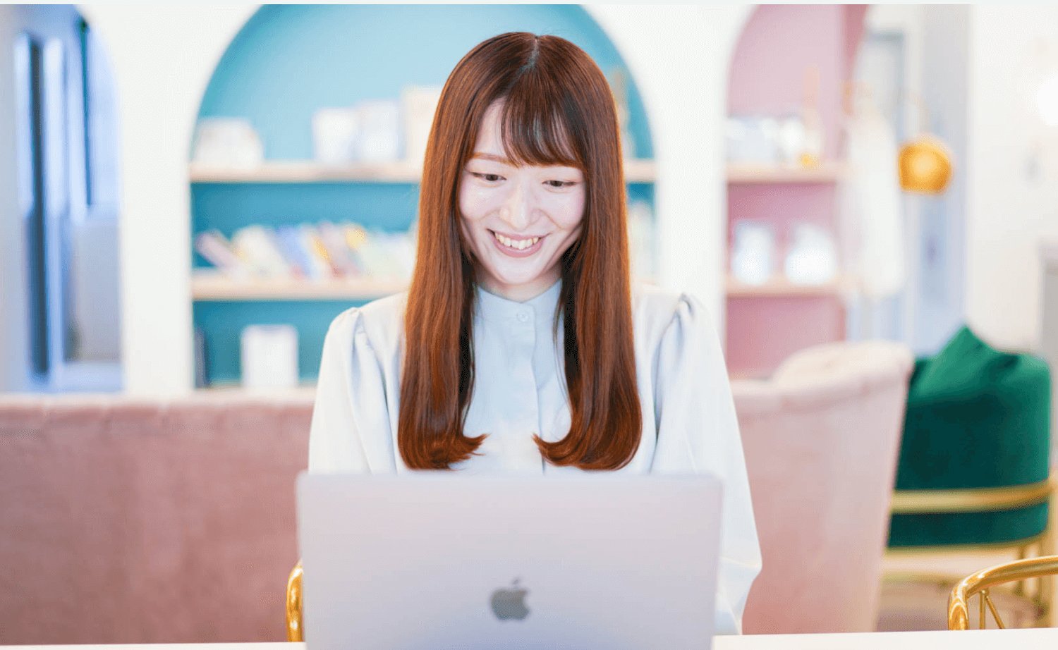 カフェの店内で、装飾された壁面の前でノートPCを操作しながら微笑む女性