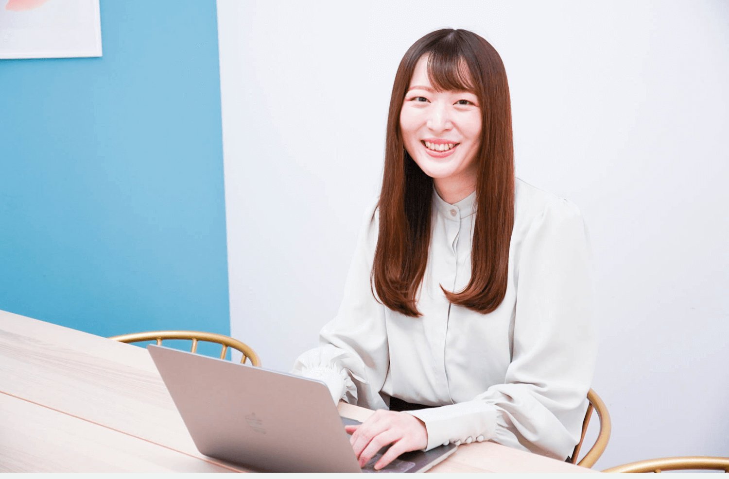 木製テーブルでノートPCを操作しながら微笑む女性。青い壁面が背景