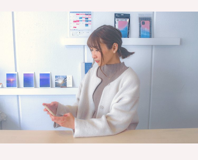白い棚の前でスマートフォンを操作する、白いニットカーディガンの女性