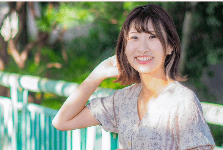 緑の街路樹と白いフェンスを背景に、髪に手を当てて微笑む花柄ブラウスの女性