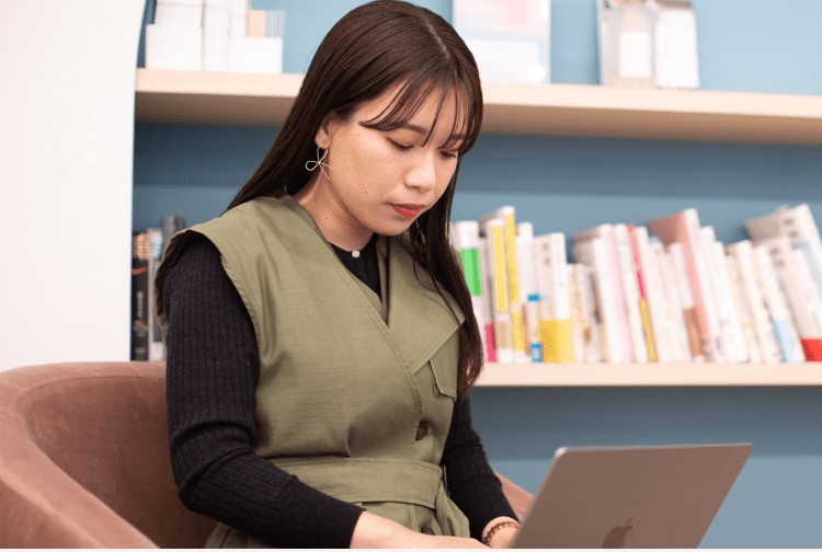 本棚を背に、ソファに座りノートPCを操作する女性