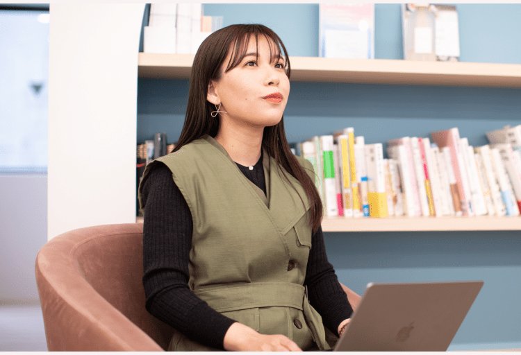 本棚を背に、ノートPCを操作しながら遠くを見上げる女性