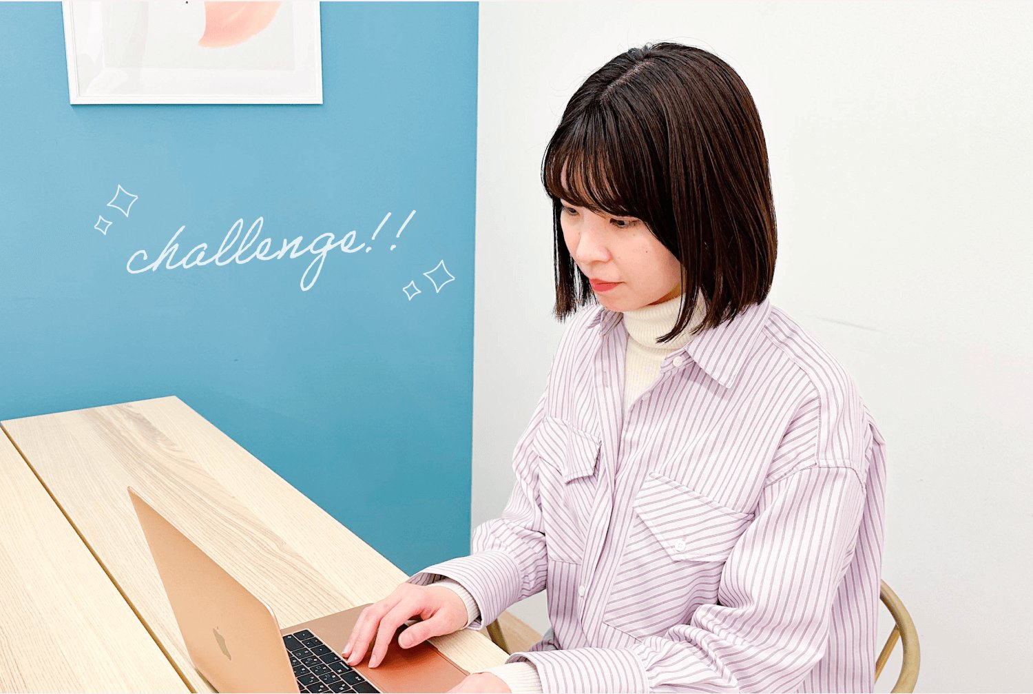 水色の壁に「challenge!!」の手書き文字とキラキラの装飾。木のテーブルでノートパソコンに向かう女性
