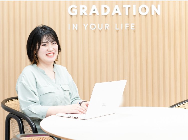 「GRADATION IN YOUR LIFE」の壁面サイネージの前でノートパソコンを開いて微笑むゆかりさん